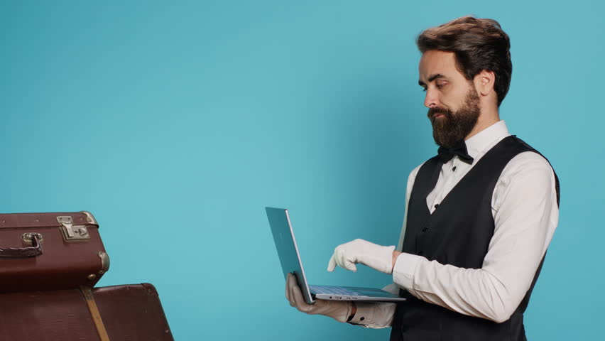 Hotel porter working on laptop and posing next to pile of luggage in studio, reviewing list of guests with online bookings. Young bellman ensuring good experience in tourism sector. Royalty-Free Stock Footage #1108410499