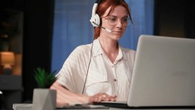call center operator, young woman with a microphone and monitors communicates via video conference on laptop with client while sitting at home at table