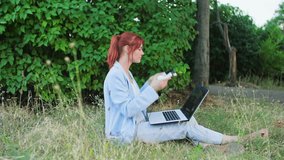 outdoor work, young female call center employee uses a headset and communicates by video link on a laptop while sitting on the grass in park