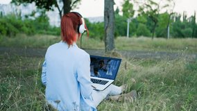 freelancing, young woman uses a headset and communicates via video link on a laptop while sitting in a park backdrop of trees