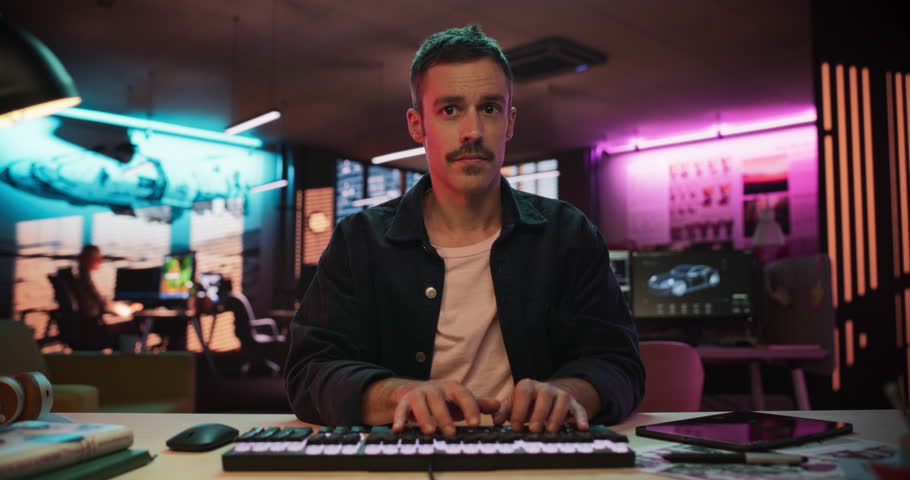 Portrait of a Male Software Developer Writing Code in a Creative Office with Neon Lights. Young Man Focused on Typing on Keyboard and Working on Desktop Computer. Point of View of Screen Monitor
