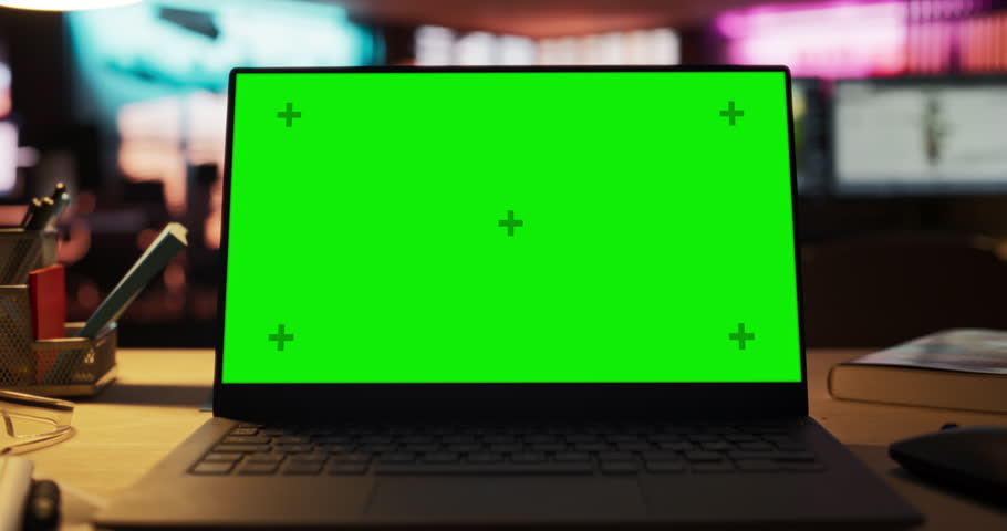 Zoom Out of Mock-up Green Screen on a Laptop Standing on the top of a Wooden Table. In the Background Stylish Modern Office Studio in the Evening with Neon Lights. Desk of Creative Designer