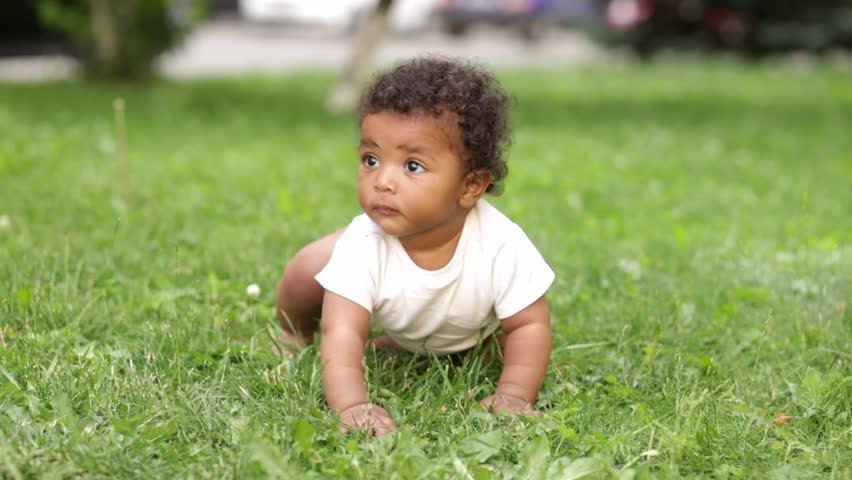 a six-month-old black African-American kid crawls on the green grass on the lawn in the summer playing with grass