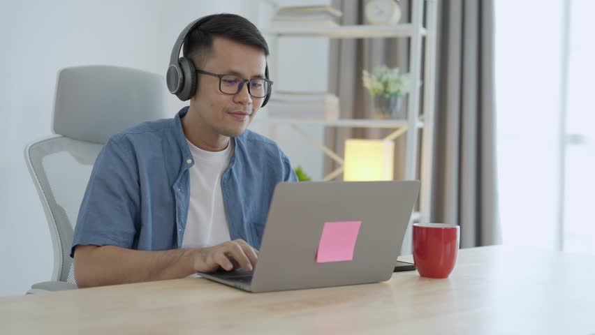 Asian freelance business man wearing headphones making video call in home office, using laptop, talking, watching webinar or studying language, speaking, online training, explaining, e-learning. Royalty-Free Stock Footage #1108419007