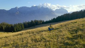AERIAL: A lady rests on a picturesque autumn alpine meadow with her adorable dog. After a hike with her furry friend, she enjoys magnificent views of mountains in embrace of unspoiled nature in fall.