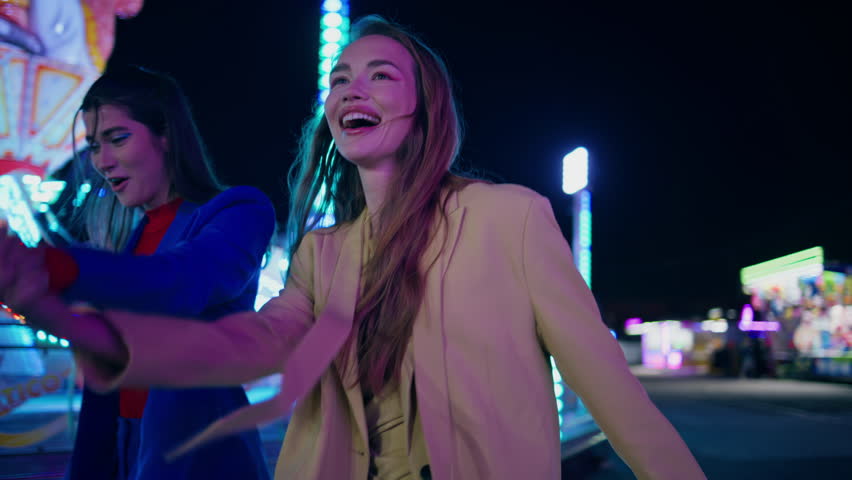 Happy girls dancing city amusement park at night. Cheerful girlfriends holding hands enjoying date weekend at illuminated carousel fair. Smiling beautiful friends couple walking having fun together