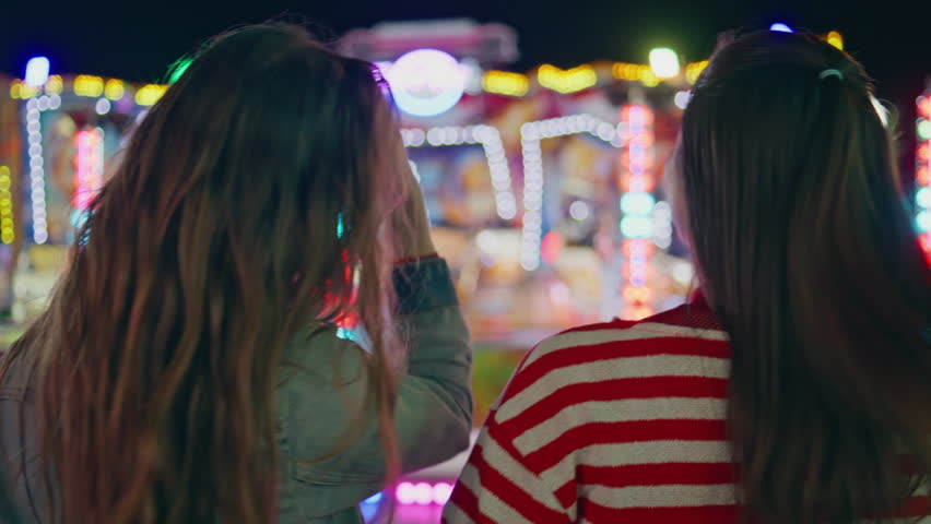 Closeup fun friends resting colourful amusement luna park. Two girls hang out watching blinking carousel at festival rear view. Relaxed joyful teenagers at city carnival. Weekend entertainment leisure Royalty-Free Stock Footage #1108427511