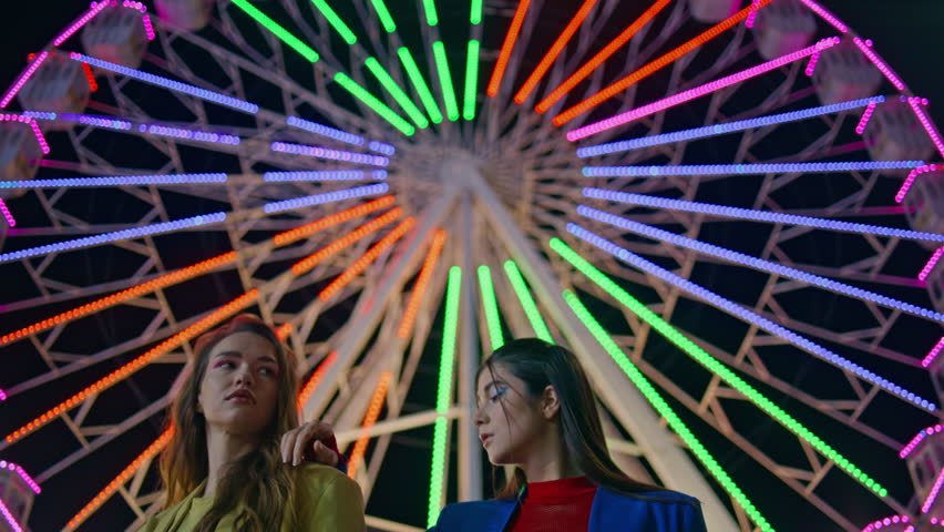 Closeup beautiful girls posing at colorful ferris wheel. Fashion models at night resting amusement luna park carousel. Two gorgeous female girlfriends lean each other looking camera at blinking lights