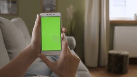 Young Woman Home Lying on a Couch with Green Screen Smartphone in Vertical Mode. Girl Using Touchscreen Mobile Phone. Girl Using Smartphone, Browsing Internet, Watching Video Content, Blogs. POV.
