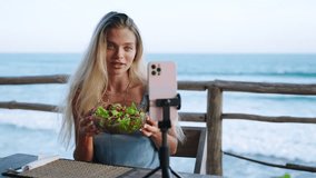 Food blogger woman films video about green salad bowl, dieting for fit skinny body at cafe, sea view. Travel influencer tells about local cuisine, diet on social network. Vegan female shoots vlog.