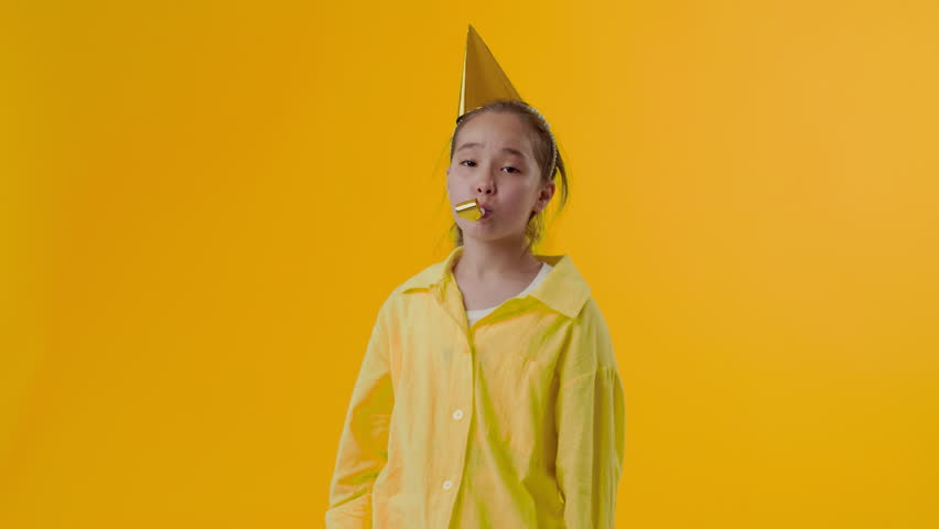 Young asian girl in the paper cap blowing a party horn and looking at the camera with an unhappy face on a yellow background. Concept of lack of interesting activities for a child at the party | Shutterstock HD Video #1108437175