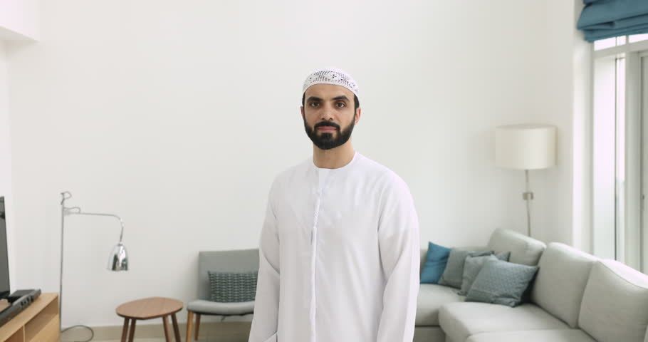 Smiling middle-aged Muslim man in traditional white kandura garment standing indoors, look at camera. Portrait of happy Arabian single male posing in modern living room. Religion, culture, traditions Royalty-Free Stock Footage #1108437743