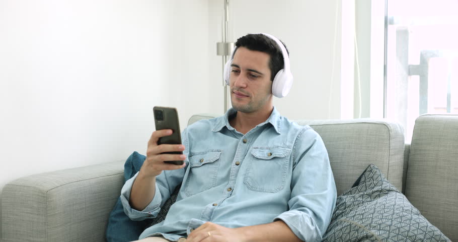 Hispanic man in wireless headphones holds smartphone choose podcast for listening use audio digital streaming media services provider resting on sofa spend leisure at home. Wireless, modern tech usage Royalty-Free Stock Footage #1108437789