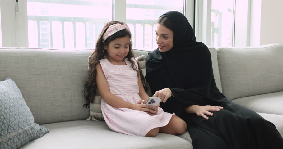 Attractive Arabian woman in traditional hijab clothing spend time on internet with little 5s adorable daughter using modern smart phone sit on sofa at home, play virtual on-line game, discuss new app Royalty-Free Stock Footage #1108437833