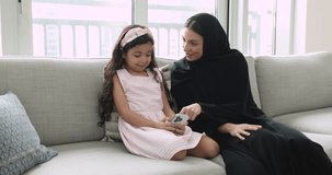 Attractive Arabian woman in traditional hijab clothing spend time on internet with little 5s adorable daughter using modern smart phone sit on sofa at home, play virtual on-line game, discuss new app