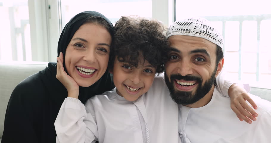 Close up portrait loving Muslim family with little 5s son in traditional wear, smile look at camera. Islamic woman wear hijab, her husband in dishdasha and kufi on head posing with kid seated on sofa Royalty-Free Stock Footage #1108437861
