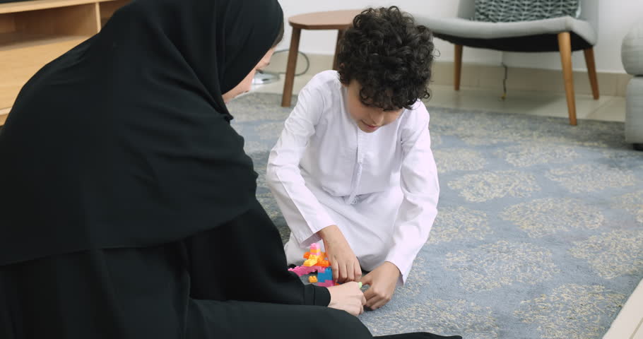 Young 30s Muslim woman in traditional clothing sit on floor with little 5s adorable son in white thobe play plastic toy set in living room. Motherhood, playtime of loving family on weekend at home Royalty-Free Stock Footage #1108437865