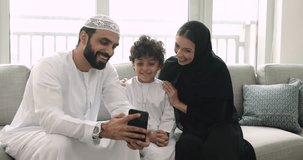 Happy Muslim family in traditional clothing using cellphone, having videoconference call with relatives, enjoy cool mobile application, have fun with new filter, play video game sit on sofa at home
