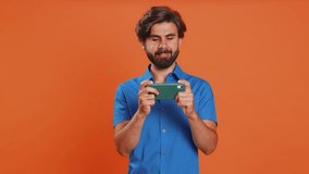 Worried funny addicted man enthusiastically playing racing video games on mobile phone. Happy middle eastern guy using smartphone gadget app with drive simulator isolated on orange studio background