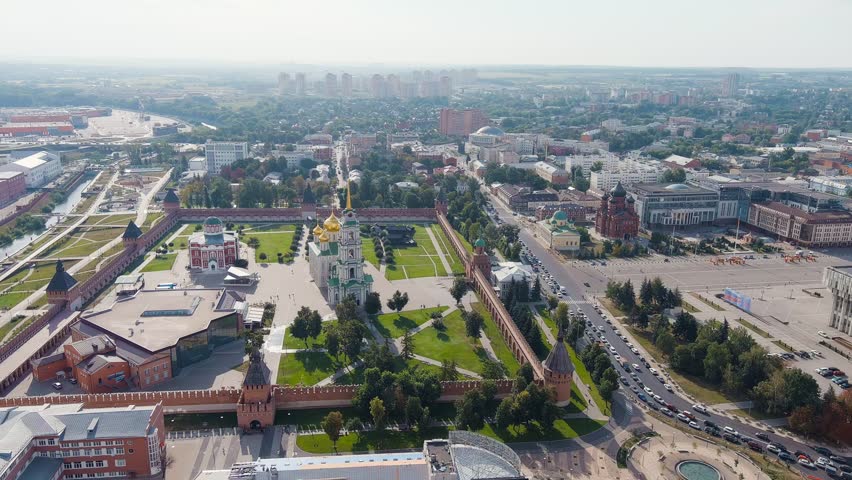 Tula, Russia. The Tula Kremlin stands as a rectangular stone fortress at the heart of the city, its red-brick defensive walls and nine towers enclosing a meticulously maintained green courtyard. At the center of the complex rises the Assumption Cathedral with its tall bell tower and gleaming golden domes, accompanied by the red-and-white Epiphany Cathedral. The fortress occupies six hectares in the historic center, surrounded by a dense urban grid of residential and administrative buildings, with a busy multi-lane road running along its eastern perimeter. The camera circles the kremlin from a high aerial perspective, revealing the full extent of the fortification walls, the geometric lawns within, and the contrast between the ancient citadel and the modern city beyond. Construction of this defensive complex began in 1507 and was completed in 1520, making it one of the oldest stone fortresses in Russia and a key link in the Great Abatis Border that once shielded Moscow