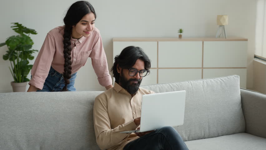 Arabian Indian couple on couch use laptop computer shopping online together pay bills order food at home choose buying internet goods give high five gesture happy man and woman approve teamwork unity Royalty-Free Stock Footage #1108447701