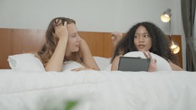 Two diverse woman on bed watching video or clip video and talking about story on digital tablet. Young girl spending time with girlfriend on weekend at home.