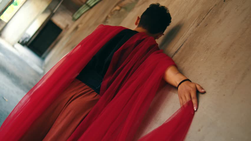 A man wearing a black shirt and rust-colored pants walks through a derelict concrete building, trailing a long, sheer red fabric. Filmed from behind with a Dutch angle, evoking a sense of mystery.