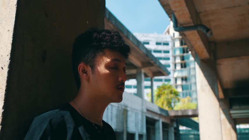 A young man with curly hair leans against a concrete pillar in a sunny, abandoned building, looking around thoughtfully before taking a deep, relaxing breath.