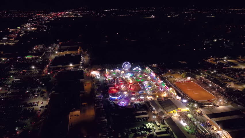 High altitude aerial establishing wide shot at night of a carnival festival fair with colorful neon lights on rides with a rodeo and large ferris wheel with crowds of people in a small town. | Shutterstock HD Video #1108457943