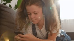 Digital fun concept. Cute smiling adolescent girl using mobile phone wearing wireless headphones, enjoying music and watching video online, lying relaxing on comfy sofa at home. Kid and gadgets