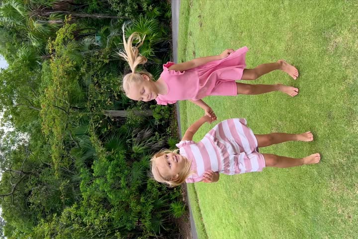 Joyful little sisters jumping holding hands on the lawn with trees in the background. Family games in park. Carefree childhood. Vertical video.