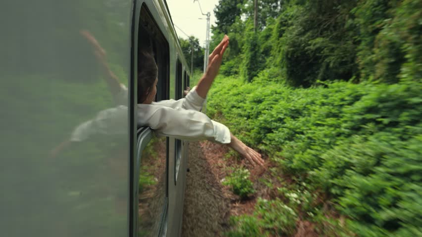 A young woman or girl puts her hands and head out from the open window of a moving train and smiles. Enjoys traveling by train, a trip to the mountains in the summer. Royalty-Free Stock Footage #1108478685