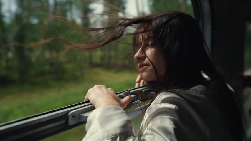 Close up view of young long-haired woman or girl looks out of an open train window while the train is moving. Hair blowing in the wind. Enjoys traveling by train, a trip to the mountains at summer day Royalty-Free Stock Footage #1108478705
