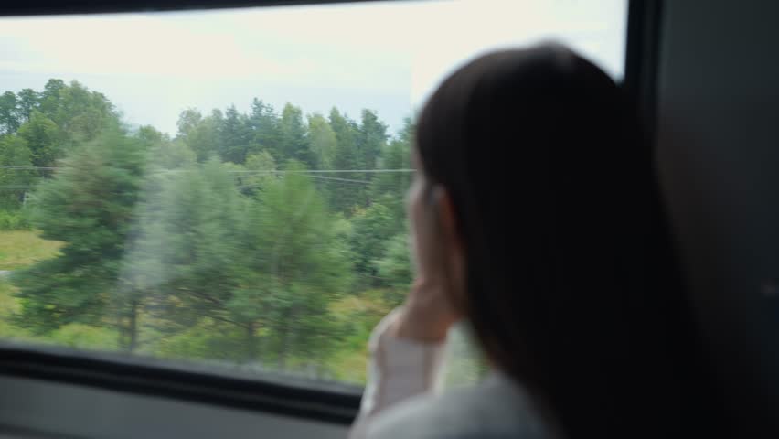 Young long-haired woman or girl close-up sitting on the passenger seat by the window in the moving train and enjoying the view from the window. Enjoys traveling by train, a trip to the mountains Royalty-Free Stock Footage #1108478721