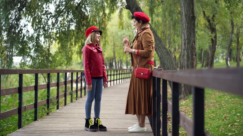 Side view wide shot positive grandmother and granddaughter talking gesturing high-five standing in autumn park on alley. Happy smiling Caucasian senior woman and teen girl enjoying weekend outdoors