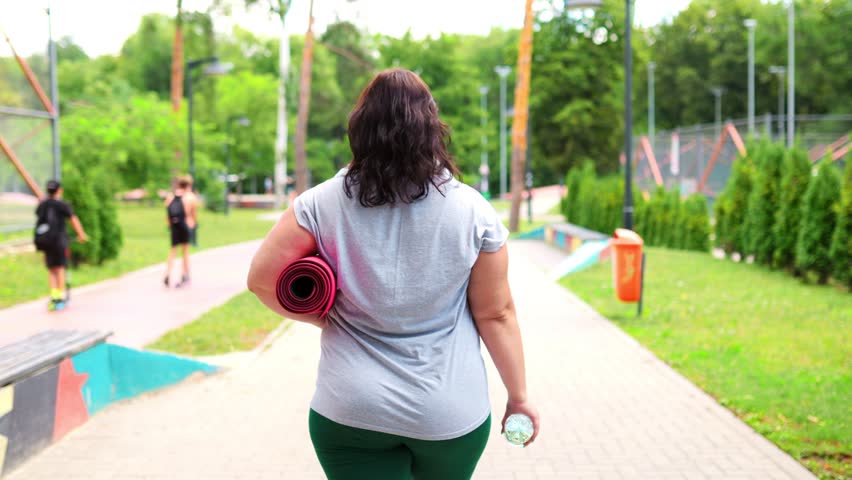 A caucasian plus size woman takes a break outdoors, rehydrating with her water bottle. It's an integral part of her weight loss regimen and healthy lifestyle. rear view Royalty-Free Stock Footage #1108490111