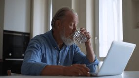 Mature older male drinking water from transparent glass having working meeting, webinar or online call at home. Happy senior aged businessman networking, communication or video conference, slow motion