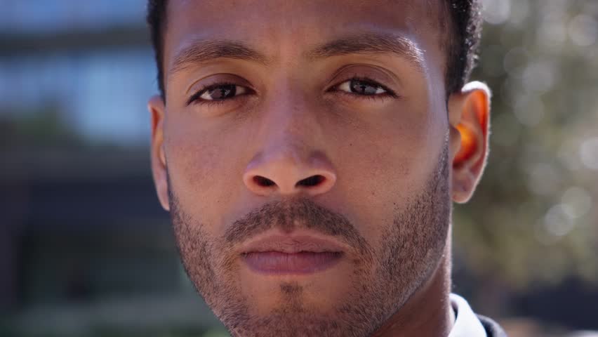 Close up portrait of young adult Latino man with serious expression. African American male looking earnest to camera outdoor with blurred background city. Millennial black boy with concerned attitude.