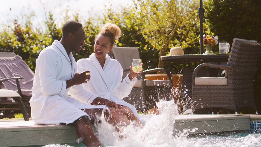 Couple wearing robes outdoors sitting and relaxing celebrating with drinks by swimming pool splashing feet in water on spa day - shot in slow motion