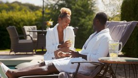 Couple wearing robes outdoors sitting and relaxing celebrating with drinks by loungers around swimming pool on spa day - shot in slow motion - Powered by Shutterstock - Get 15% off with code: PIKWIZARD15