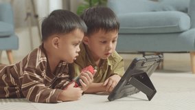 Close Up Of Asian Kids With Plastic Toy Brick Lying On The Floor Watching Movie On A Tablet Together At Home

