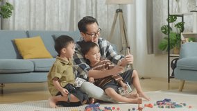Full Body Of Asian Father And Sons Playing The Construction Set Colorful Plastic Toy Brick At Home. Using Smartphone, Looking At Video Laughing Together

