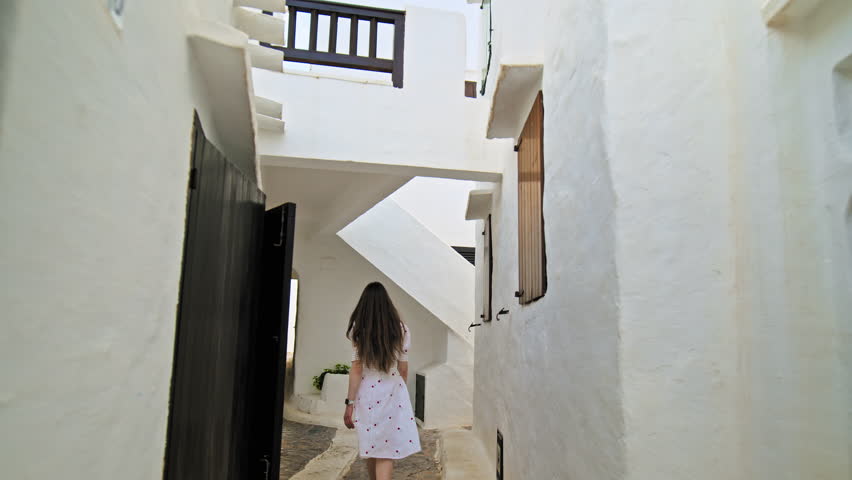 A girl tourist exploring a town with whitewashed houses and narrow streets. A young woman in a white dress walks and enjoys a small holiday village resort with tiny steps and balconies.