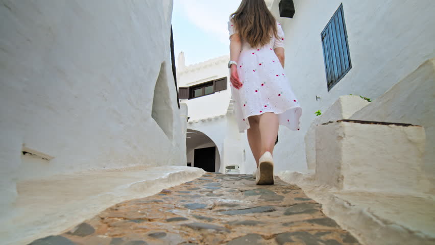 A girl tourist exploring a town with whitewashed houses and narrow streets. A young woman in a white dress walks and enjoys a small holiday village resort with tiny steps and balconies.