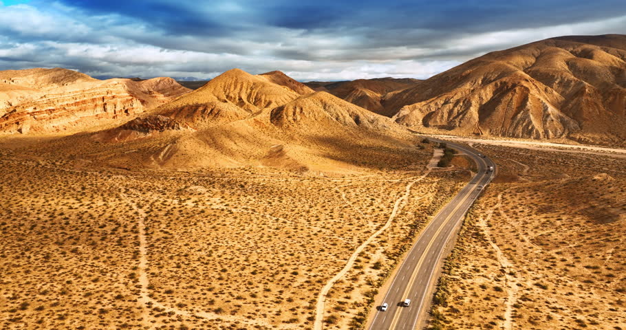 Cars going quickly by the highway among the mountains. Heavy clouds throwing shadows on the dry land. Death Valley scenery.