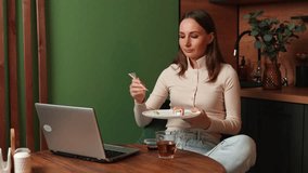 Nice brunette woman dressed in a beige sweatshirt is sitting in the kitchen and eating sushi, watching movies on a laptop. A woman rests on weekends, holidays.