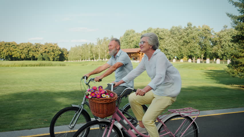 Beautiful aged husband and wife in stylish attire using vintage bikes for carefree riding on fresh air. Two cheerful people on retirement actively spending daytime at public city park. Royalty-Free Stock Footage #1108513761