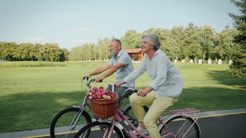Beautiful aged husband and wife in stylish attire using vintage bikes for carefree riding on fresh air. Two cheerful people on retirement actively spending daytime at public city park. Video Stok