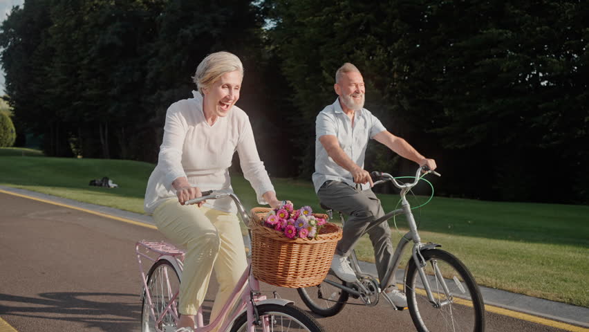 Healthy and fit elder caucasian couple riding bicycles in public park during summertime. Vibrant active seniors dressed in casual outfits having fun and raising legs while cycling. Royalty-Free Stock Footage #1108513917