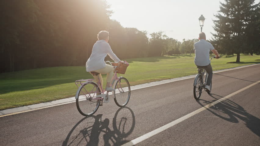 Back view of dynamic and lively aged couple cycling joyfully in public park during sunny season. Funky active man riding in front and lifting legs from pedals. Concept of fun and positive emotions. Royalty-Free Stock Footage #1108513919